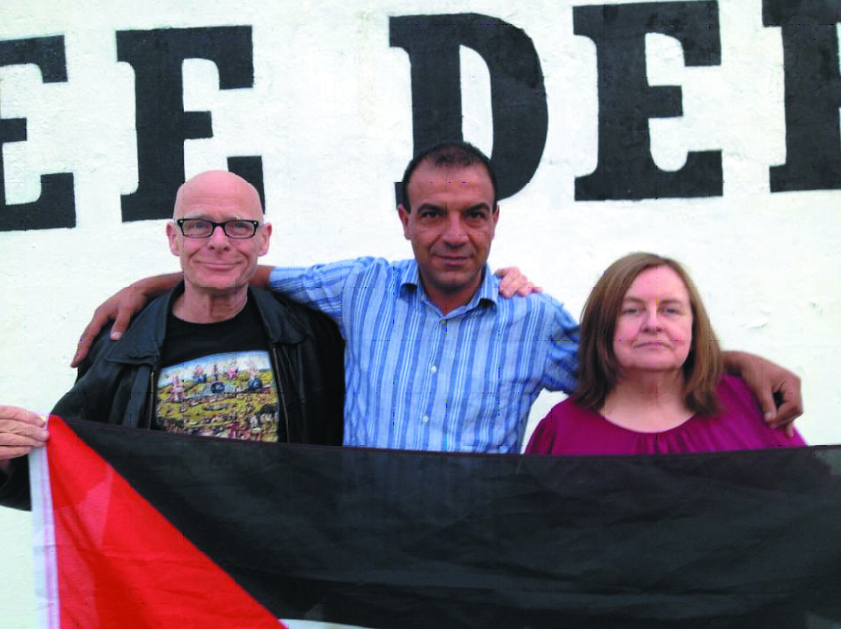 : Eamonn McCann, Bernadette Devlin, Salaf Ajarama Derry şehrinin ünlü “Şu anda Özerk Derry’ye girmekteseniz” yazılı duvarı önünde Filistin bayrağıyla.