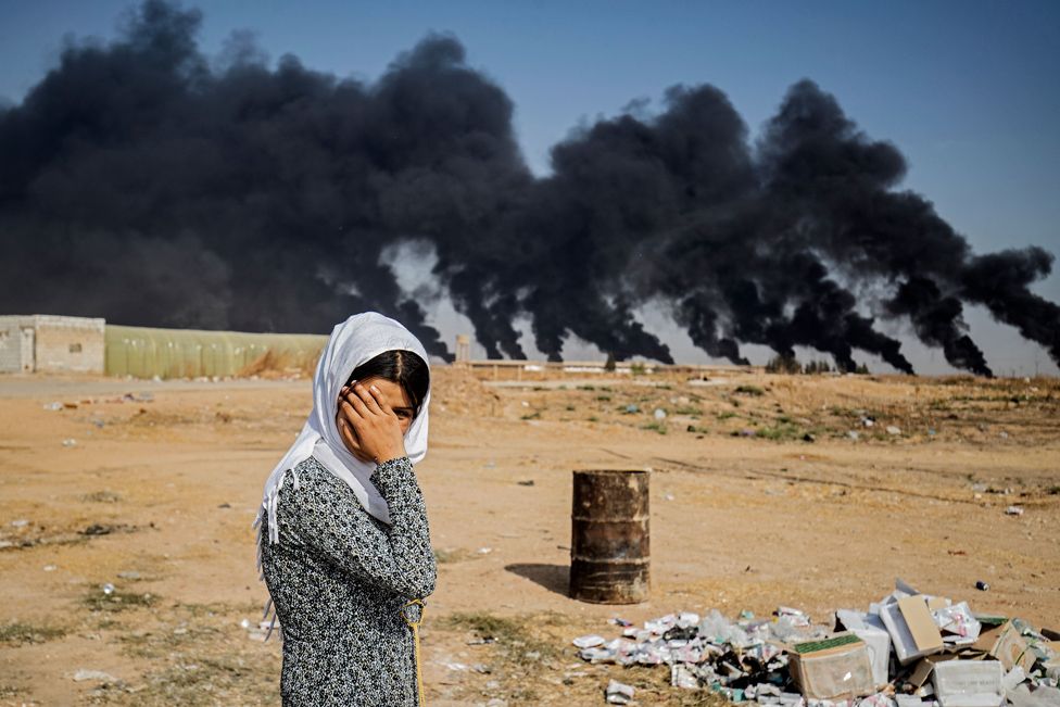 
                    A woman covers her face by a road near the Syrian Kurdish town of Ras al-Ain, along the border with Turkey. Behind her, tyre fires have been lit to decrease visibility for Turkish warplanes.
                